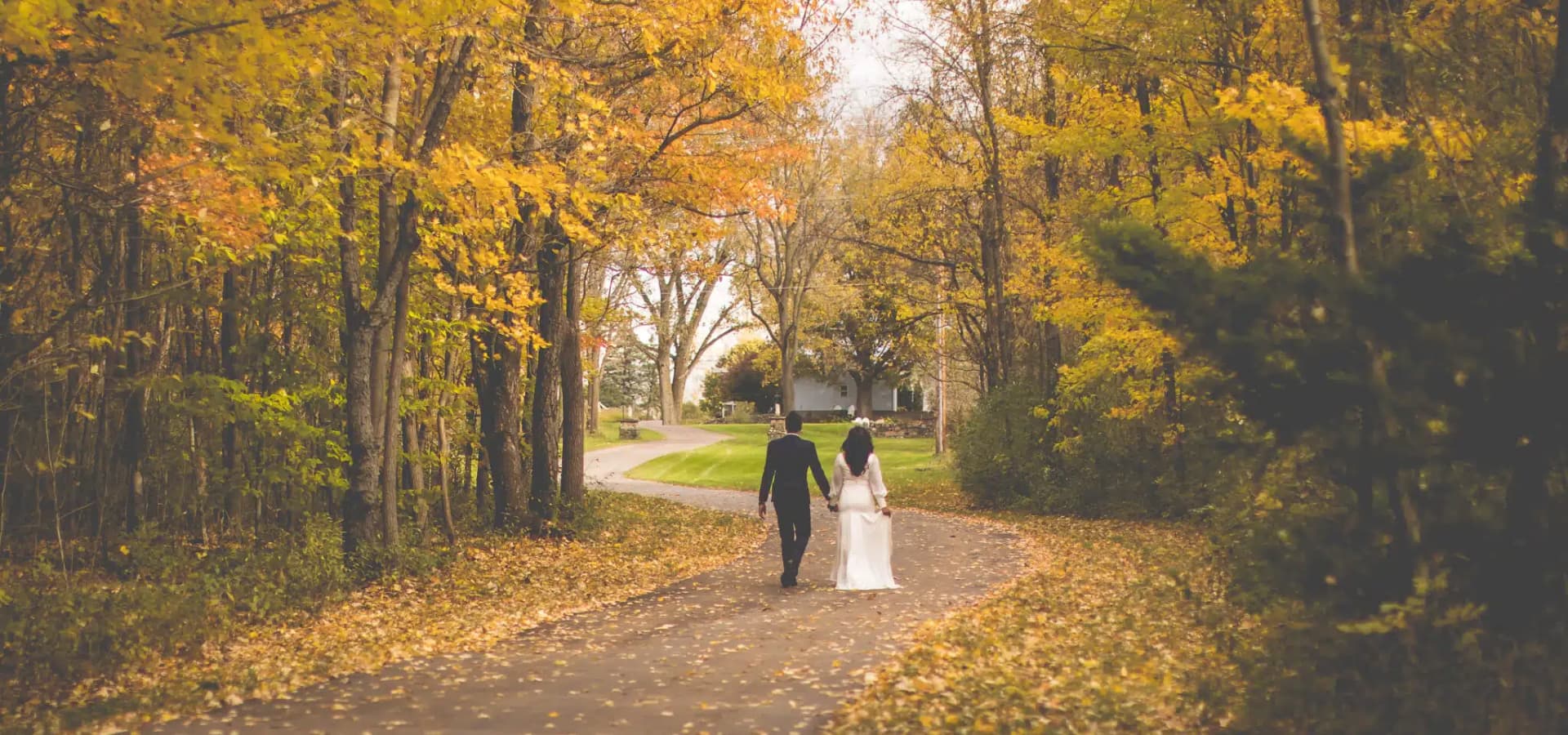 A couple walks hand-in-hand along a leaf-covered path surrounded by vibrant autumn foliage.