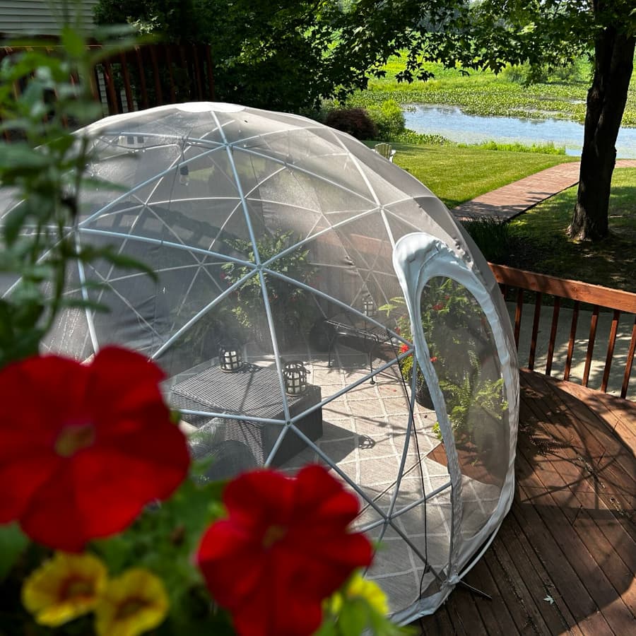 A transparent geodesic dome sits on a wooden deck, surrounded by colorful flowers with a serene pond view in the background.