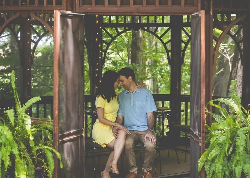 A couple sits closely together in a gazebo surrounded by greenery.