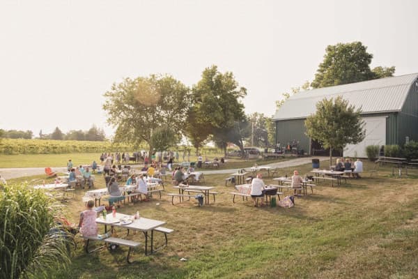 A gathering of people seated at picnic tables in a grassy area near a barn, surrounded by trees and open fields.