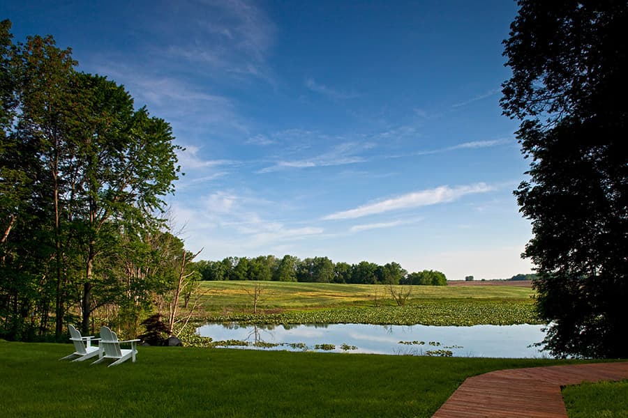 A serene landscape featuring a pond, lush greenery, and wooden chairs overlooking the water.