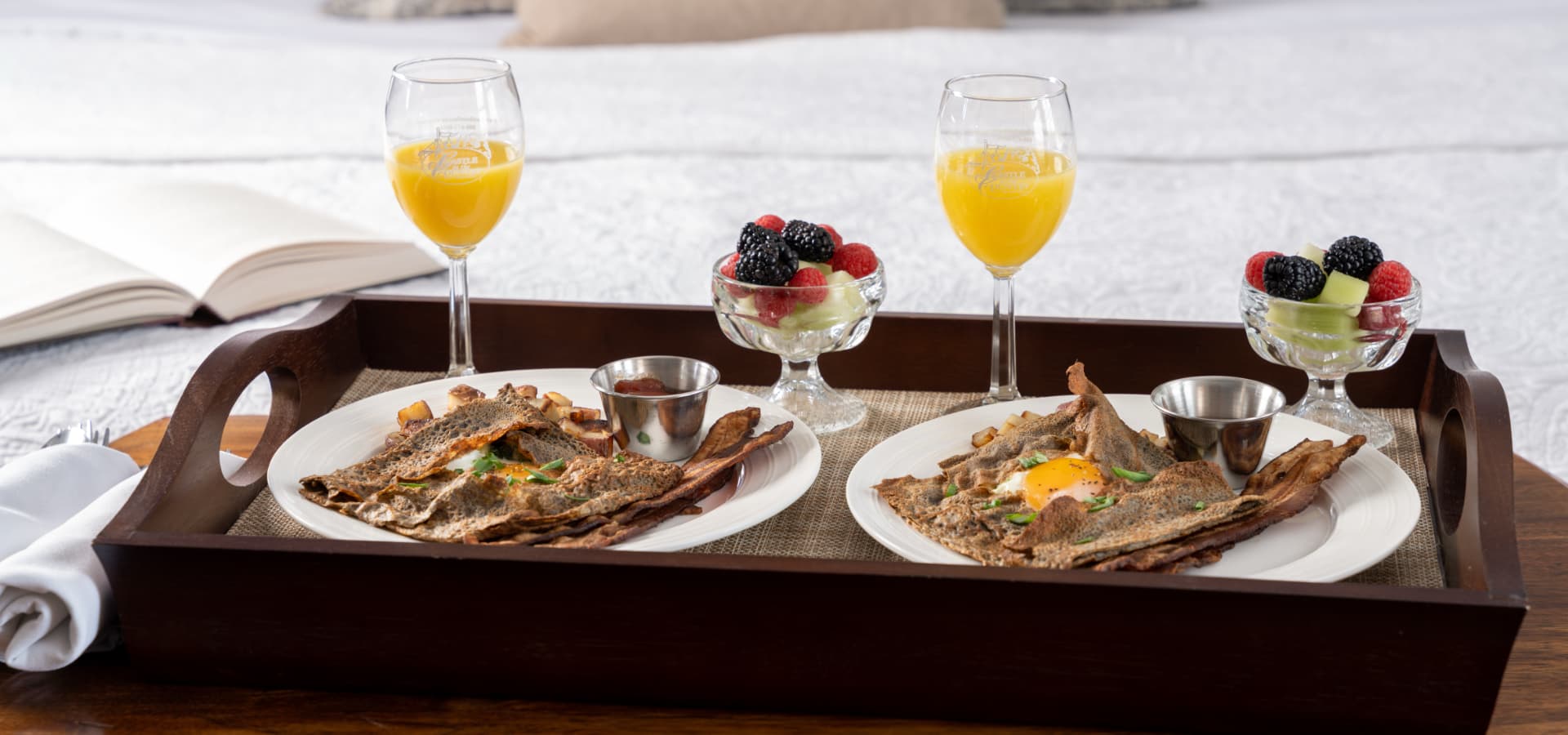 A elegantly arranged breakfast tray with crepes, fruit, and mimosas on a linen-covered table.