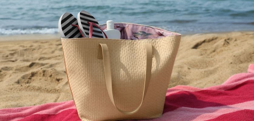A beach bag with flip-flops and a bottle sits on a sandy shore beside a red and pink striped towel.