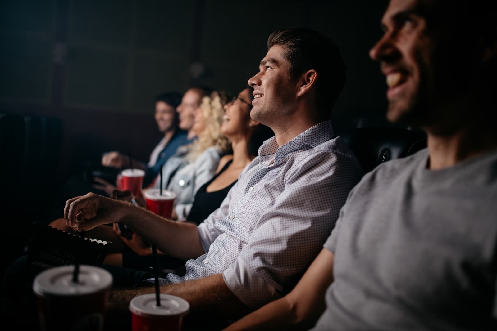 A group of friends enjoys a movie together in a theater, laughing and holding drinks.