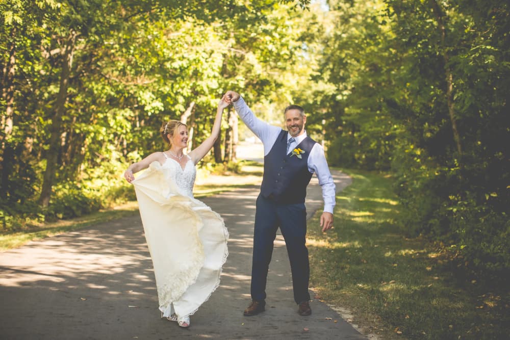 A joyful bride and groom dance along a tree-lined path, celebrating their wedding day.