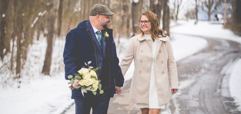 A groom and bride walk hand in hand along a snowy path, smiling at each other.