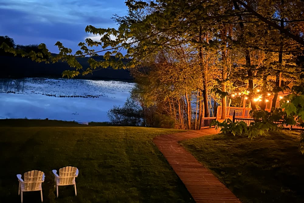 A serene lakeside scene at dusk with two white chairs and a wooden path leading to a deck adorned with string lights.