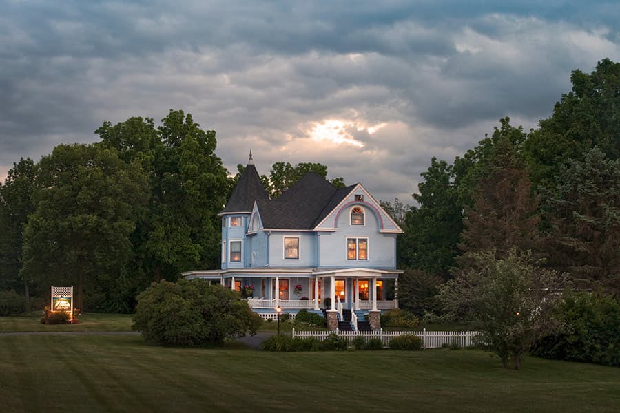 A blue Victorian-style house stands amidst lush greenery under a partly cloudy sky.