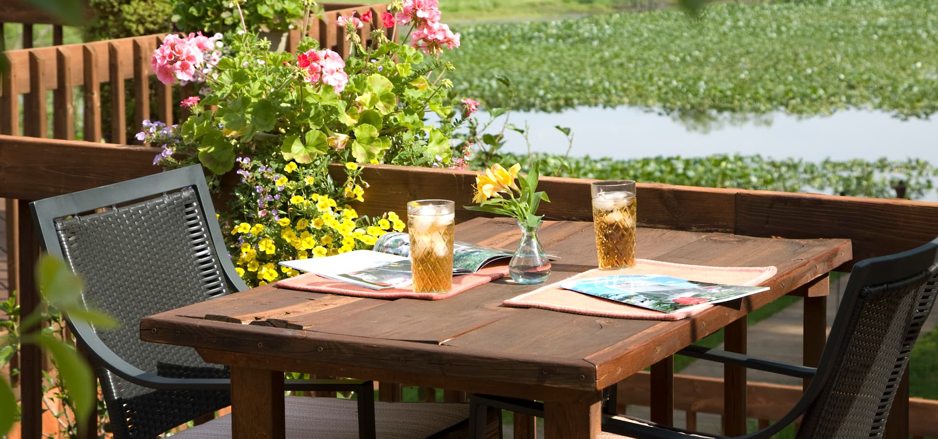A wooden table set for two with drinks, flowers, and magazines, surrounded by colorful flowers and a view of a serene waterway.