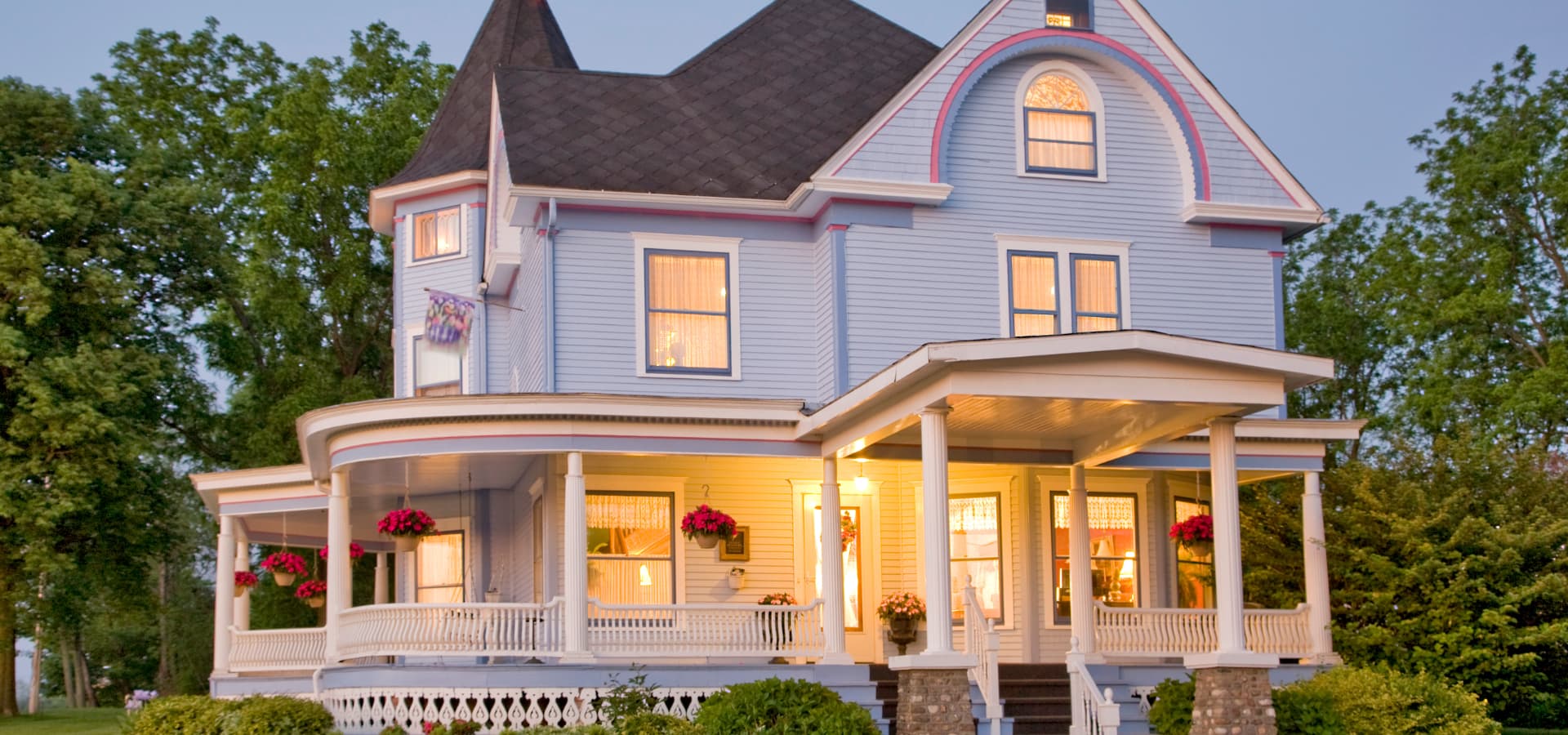A charming blue Victorian house with a bay window and a wraparound porch, surrounded by greenery.