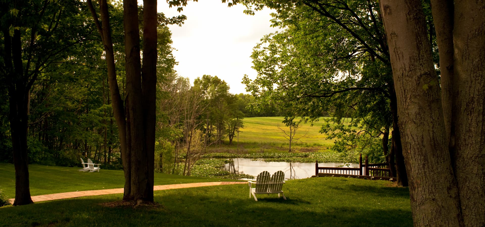 A serene landscape featuring a grassy area with two white chairs facing a calm pond surrounded by trees.