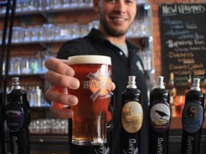 A bartender smiles while holding up a pint of beer in front of various tap handles.