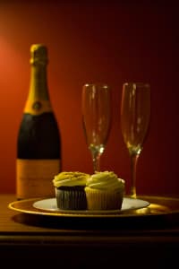 A bottle of champagne and two flutes next to a plate of cupcakes against a warm red background.