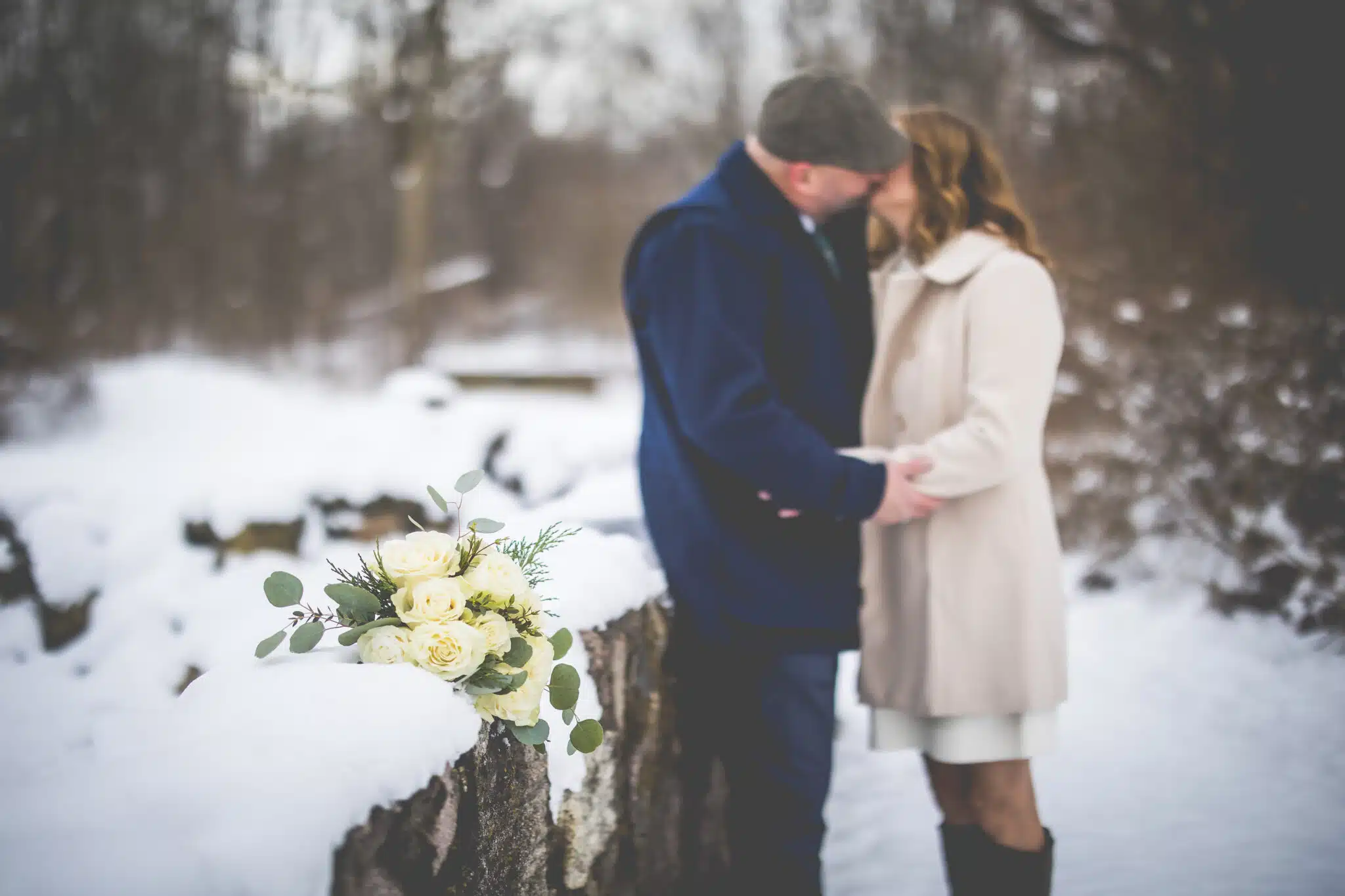 A couple kisses romantically on a snowy path, with a bouquet of white roses nearby.