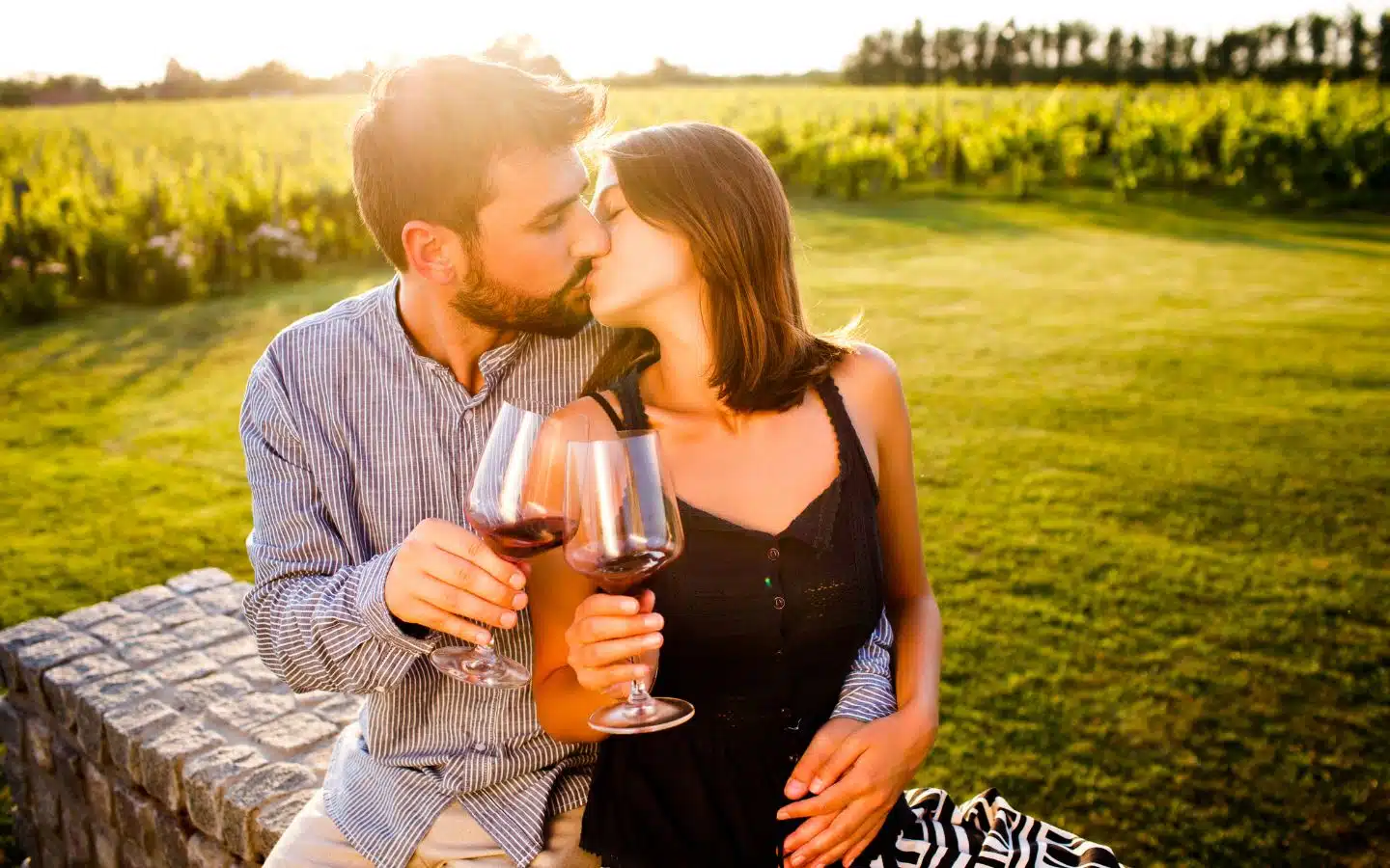 A couple leans in for a kiss while holding glasses of red wine in a sunny vineyard setting.