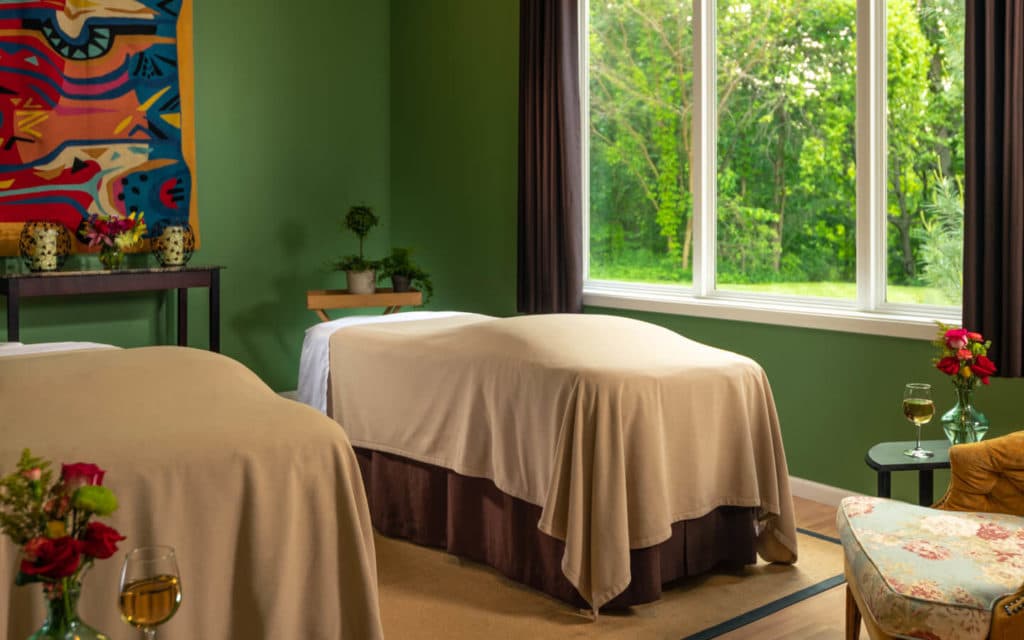 A tranquil spa room features two massage tables with beige coverings beside a large window overlooking greenery.