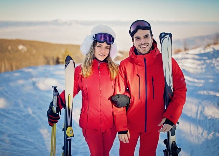 A young couple in matching red ski outfits poses with their skis on a snowy mountain.