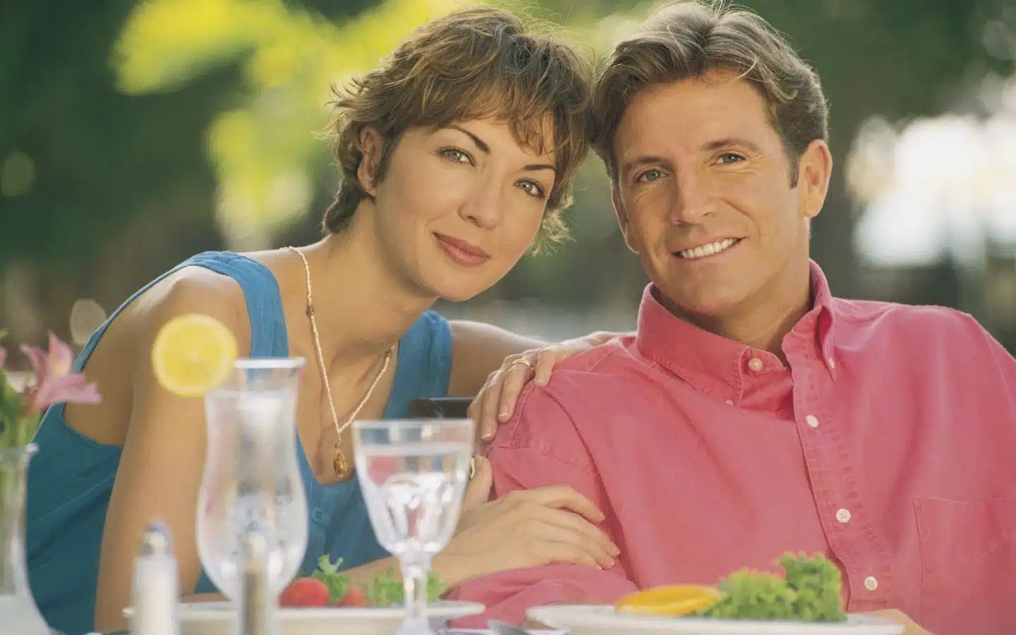 A couple smiles together at an outdoor table, surrounded by food and drinks. A couple smiles together at an outdoor table, surrounded by food and drinks.