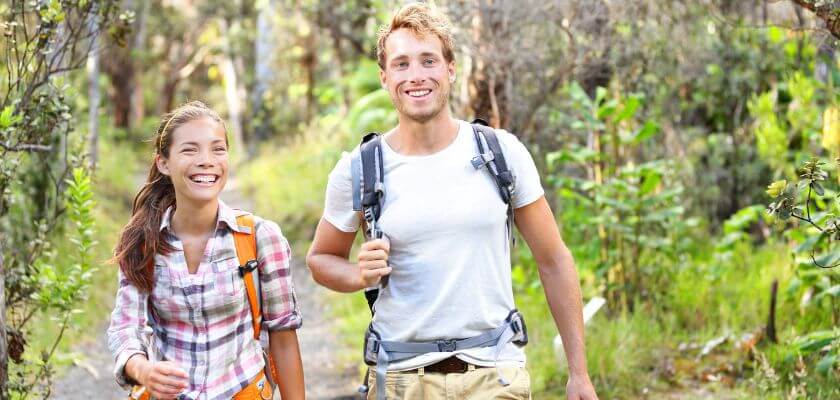 Two hikers smiling while walking on a trail surrounded by greenery.