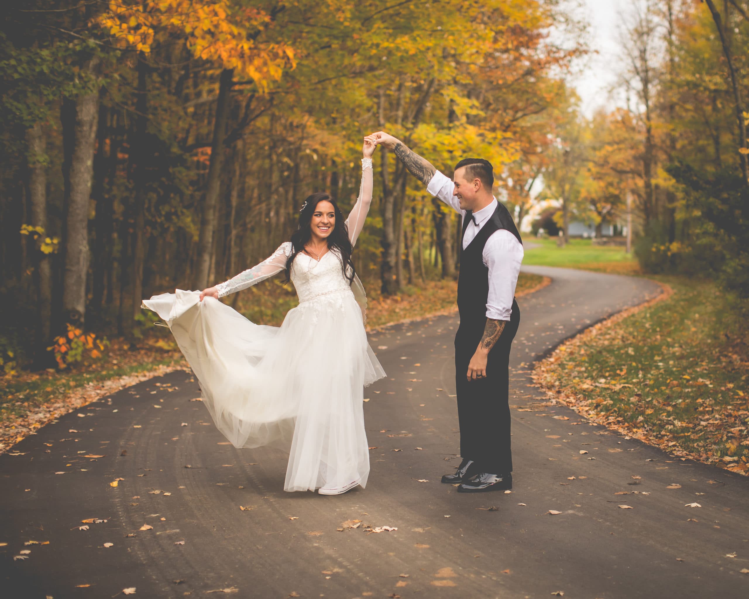 A bride and groom joyfully dance on a leaf-strewn path surrounded by autumn trees.