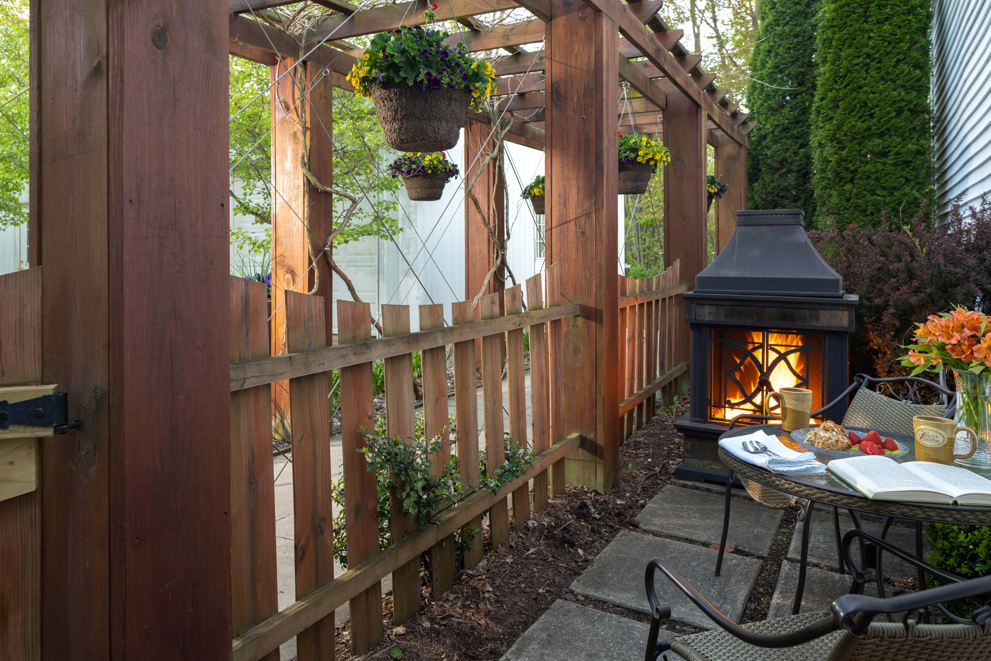 A cozy outdoor space featuring a wooden trellis, potted plants, and a small fireplace beside a table set for dining.