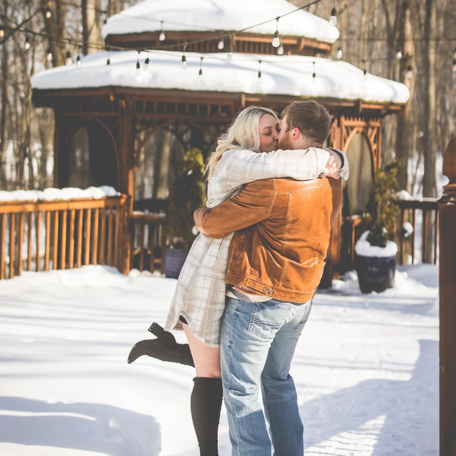 A couple joyfully kisses while embracing in a snowy outdoor setting near a gazebo.