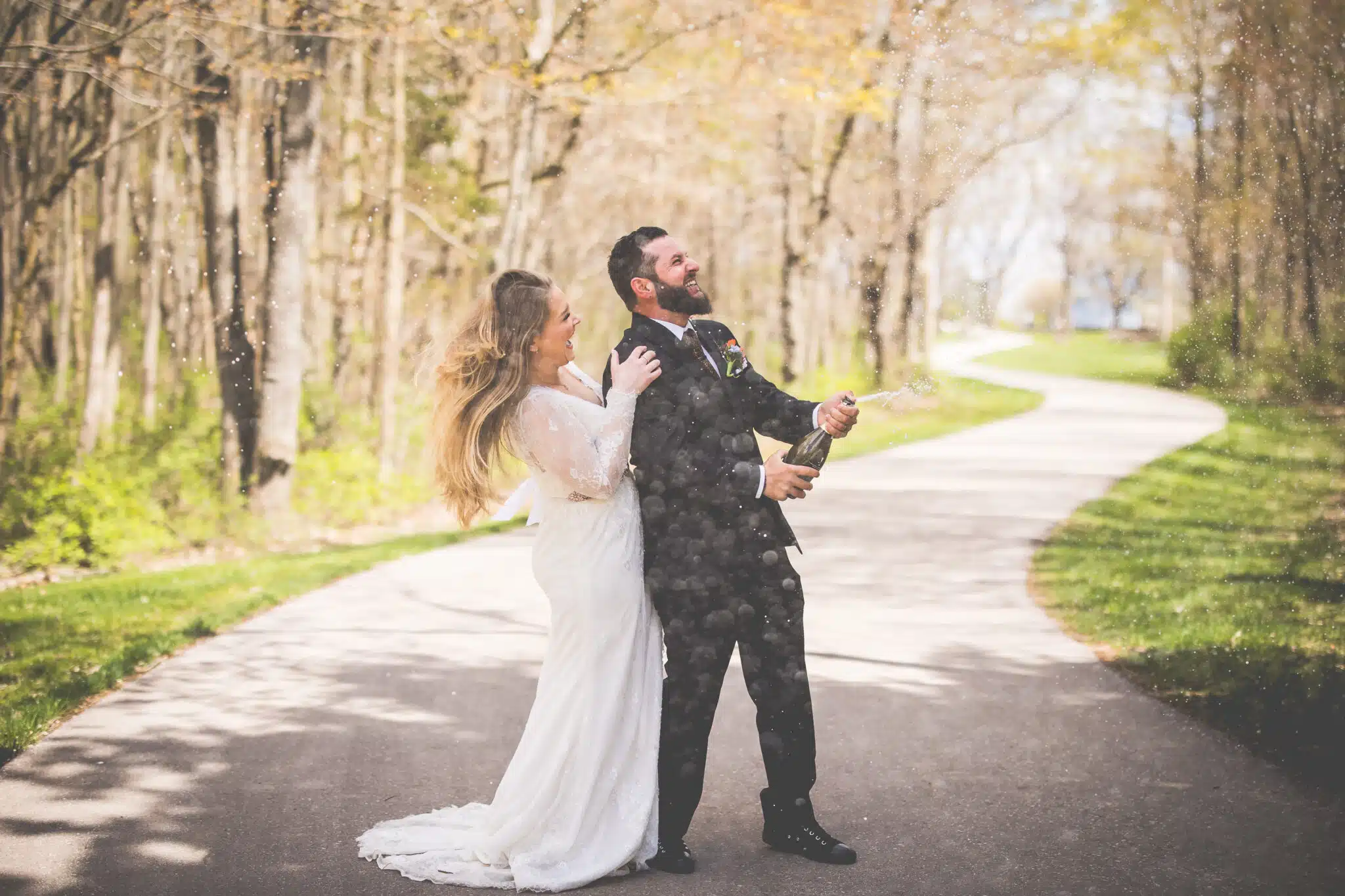 A joyful couple celebrates by popping a champagne bottle on a picturesque path in a wooded area.
