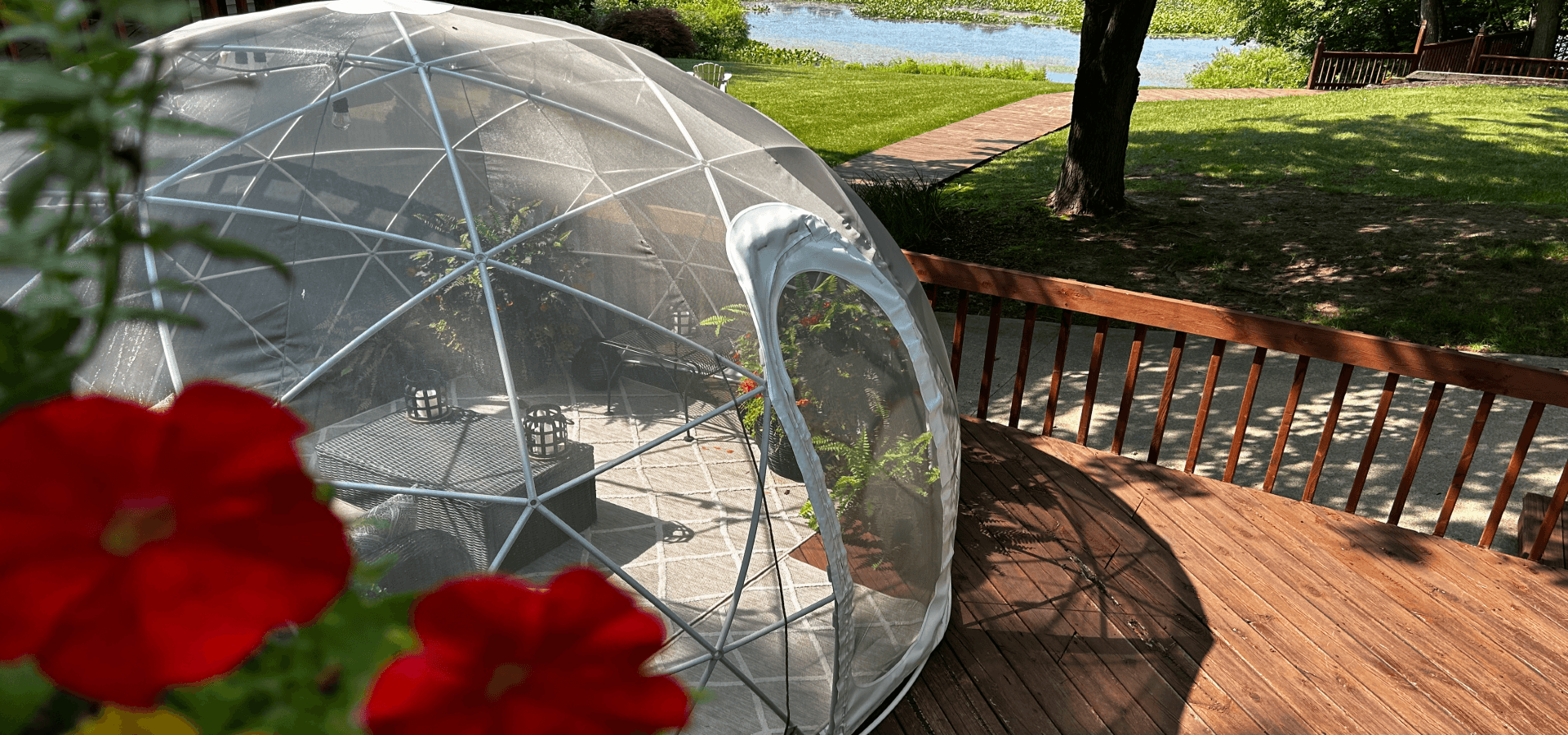 A clear geodesic dome sits on a wooden deck overlooking a serene water view, surrounded by vibrant red flowers.