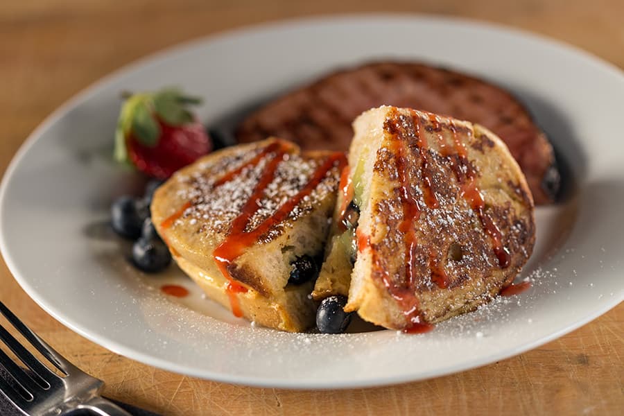 A plate of stuffed French toast drizzled with syrup, accompanied by blueberries, a strawberry, and a slice of sausage.