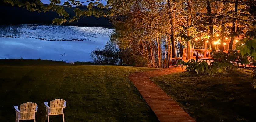 A serene lakeside scene at twilight with a path leading to a cozy porch and two empty white chairs.