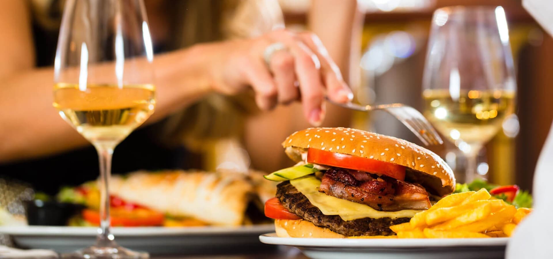 A close-up of a gourmet burger with bacon, cheese, and vegetables, accompanied by a glass of white wine and fries.