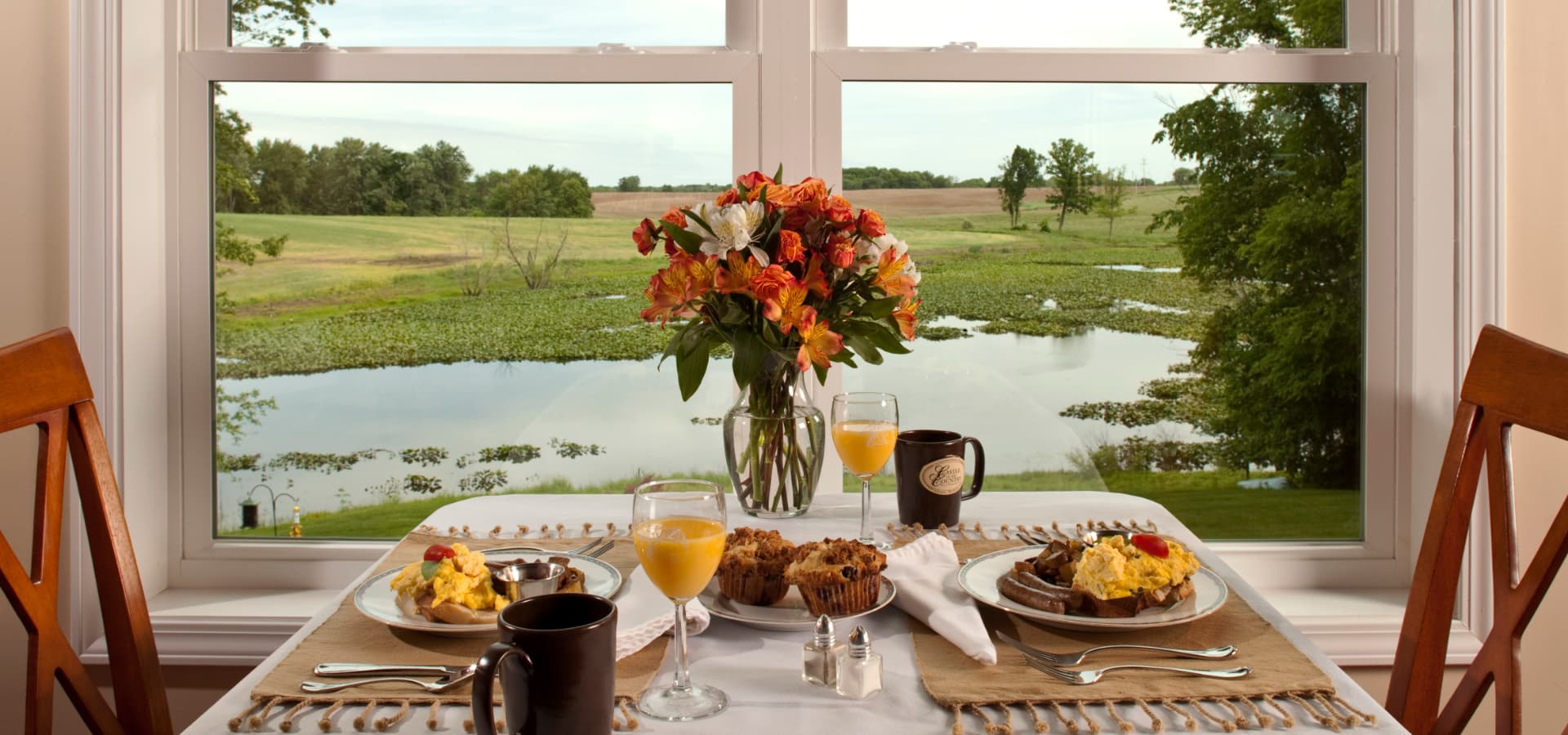 A beautifully set breakfast table featuring eggs, muffins, and a vase of flowers, with a scenic view of a pond and greenery outside.