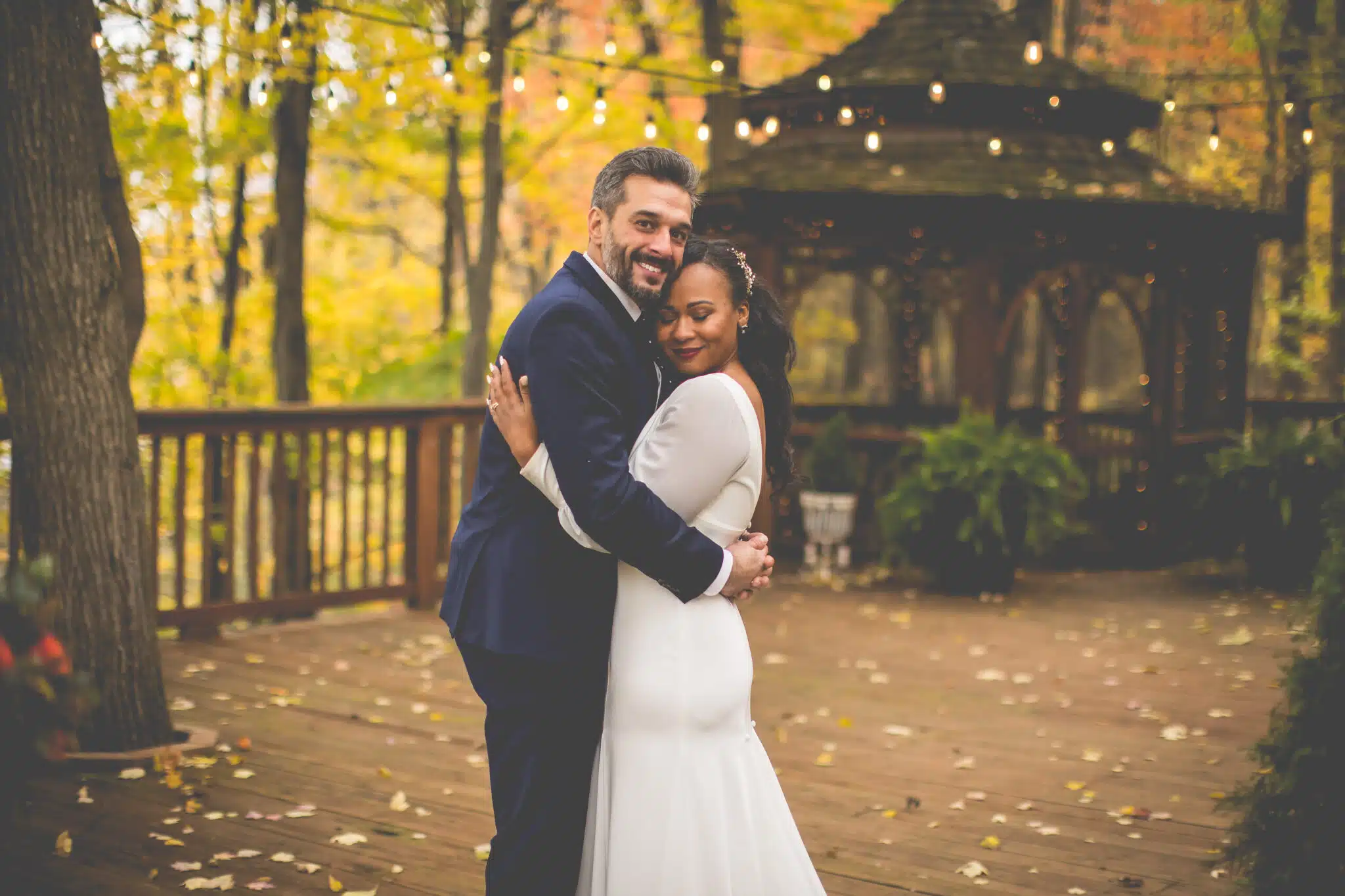 A couple embraces joyfully on a wooden deck surrounded by autumn foliage.