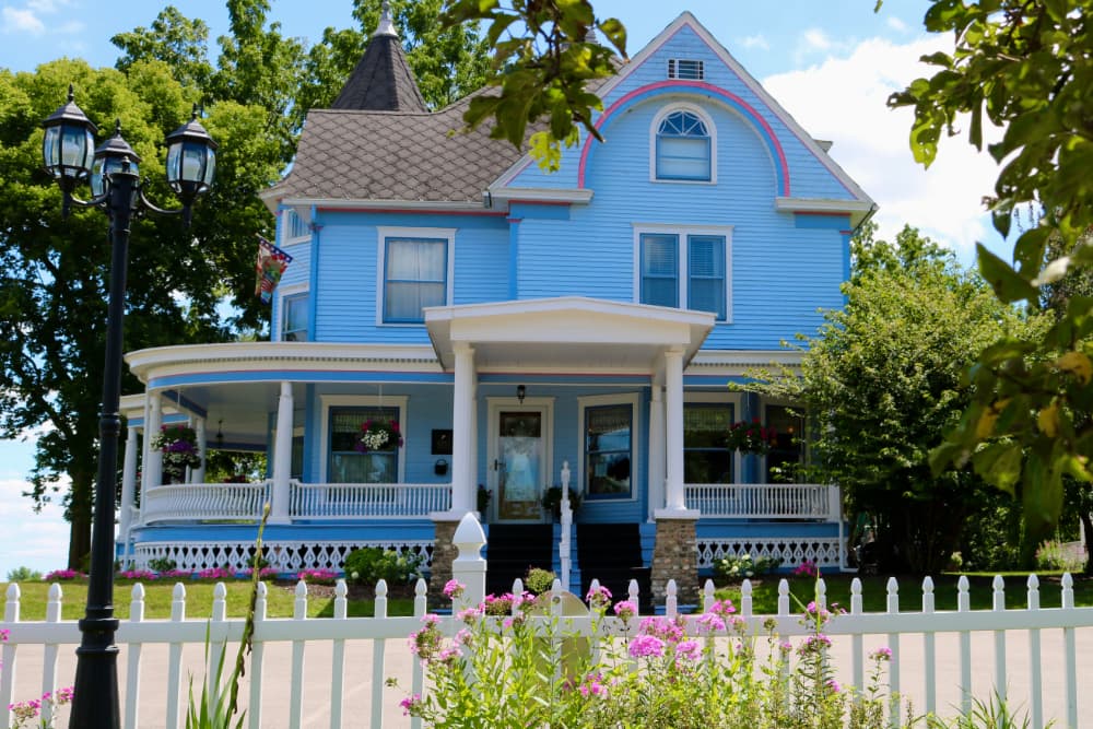 A charming blue Victorian house with a porch and colorful flower beds, set against a backdrop of trees.