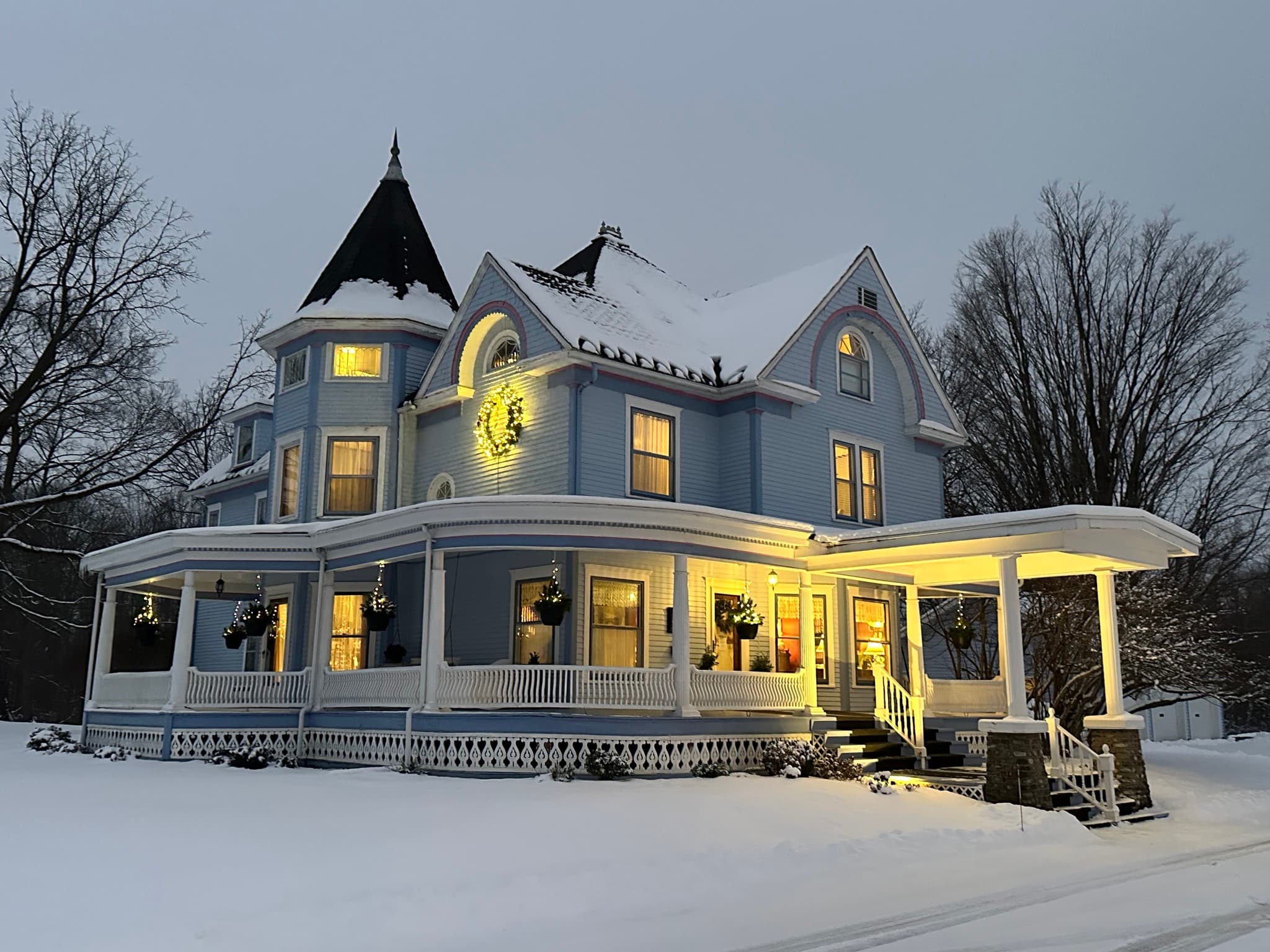 A large blue Victorian house with snow-covered grounds, illuminated warmly at dusk.