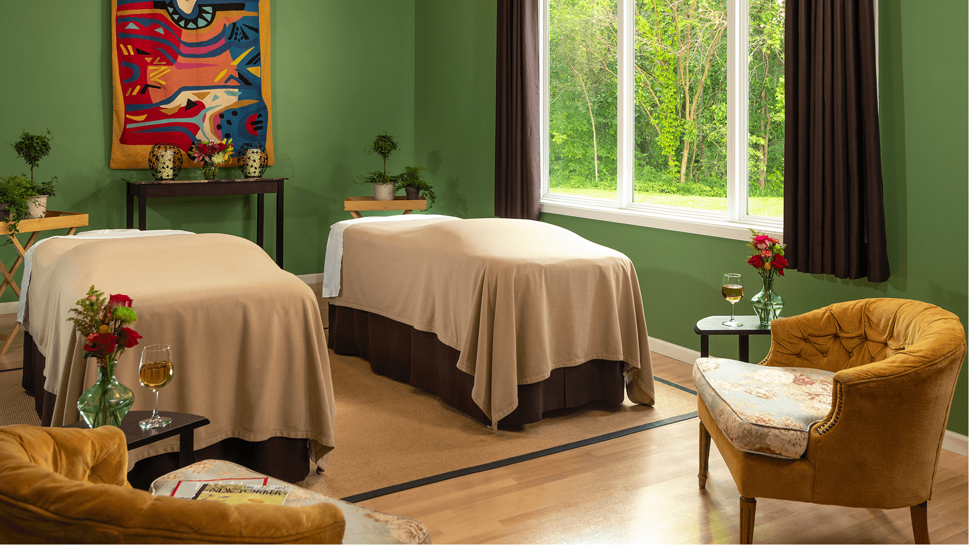 A tranquil spa room featuring two massage tables, a cozy chair, and large windows overlooking greenery.