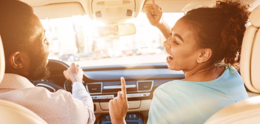 A man and woman are joyfully singing and dancing in a car, with sunlight streaming through the windows.