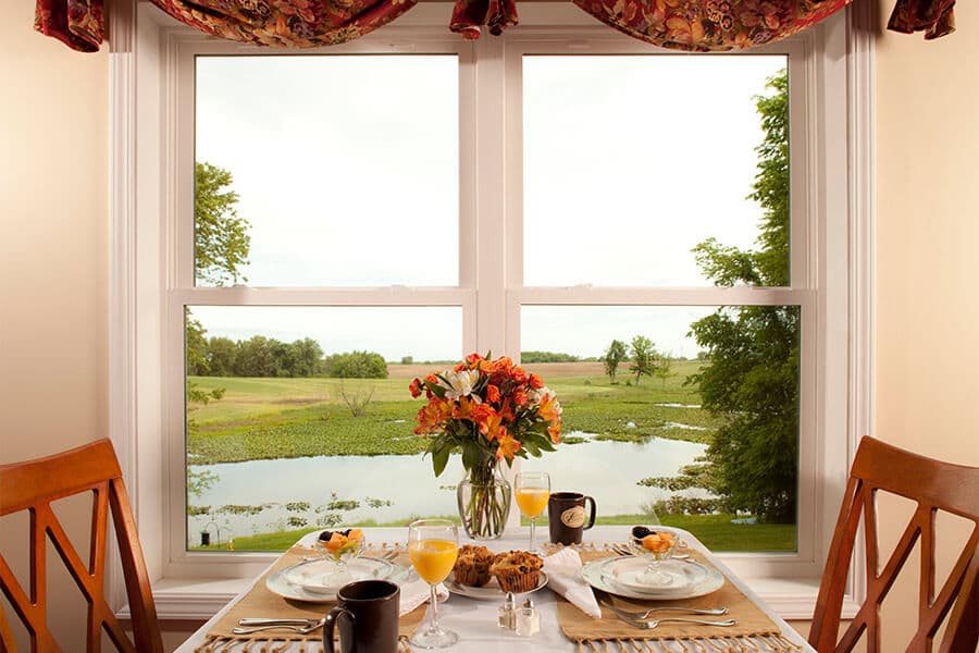 A beautifully set breakfast table with flowers and drinks overlooks a serene lake through a large window.