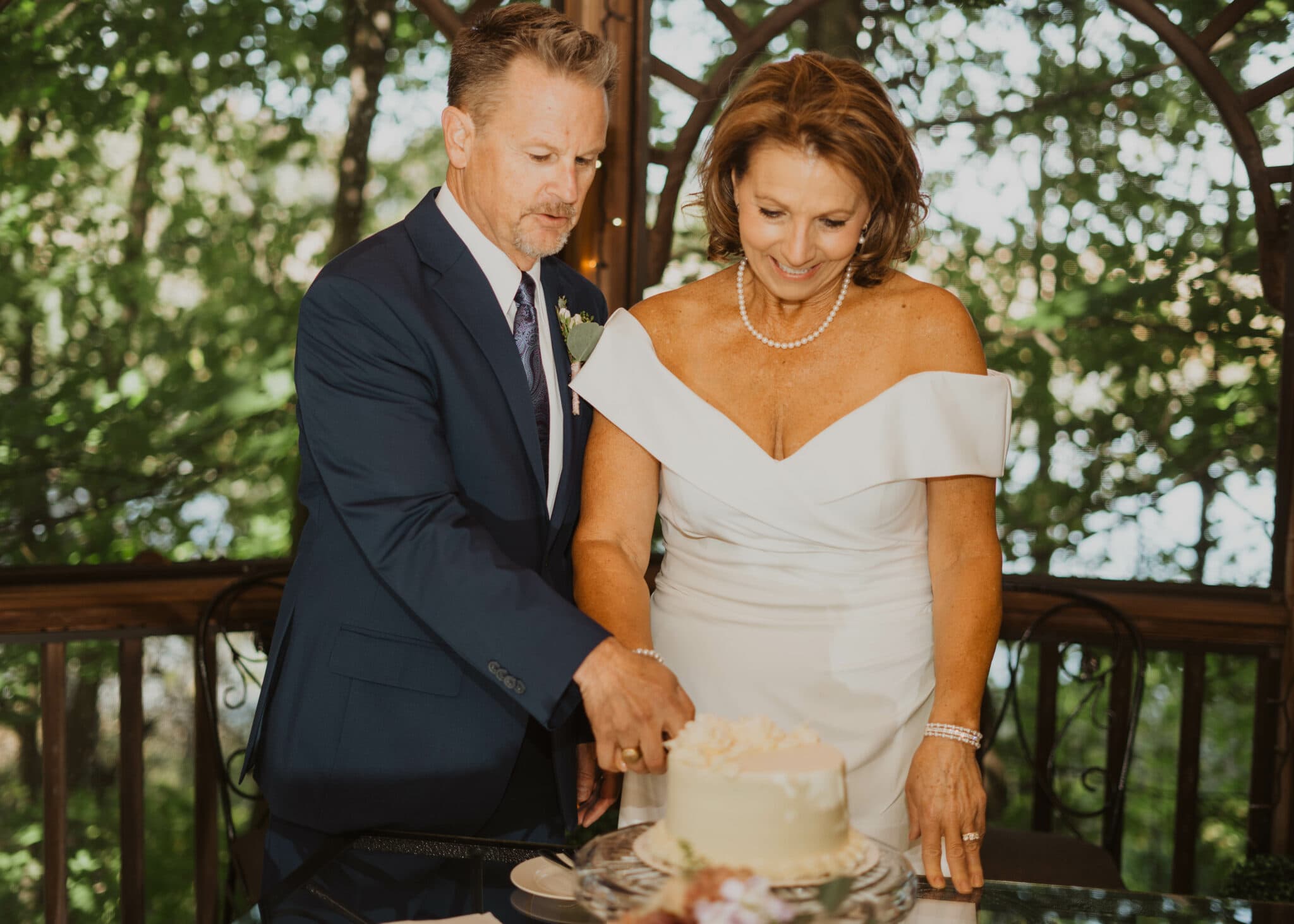 A couple joyfully cuts a wedding cake in a lush, outdoor setting.