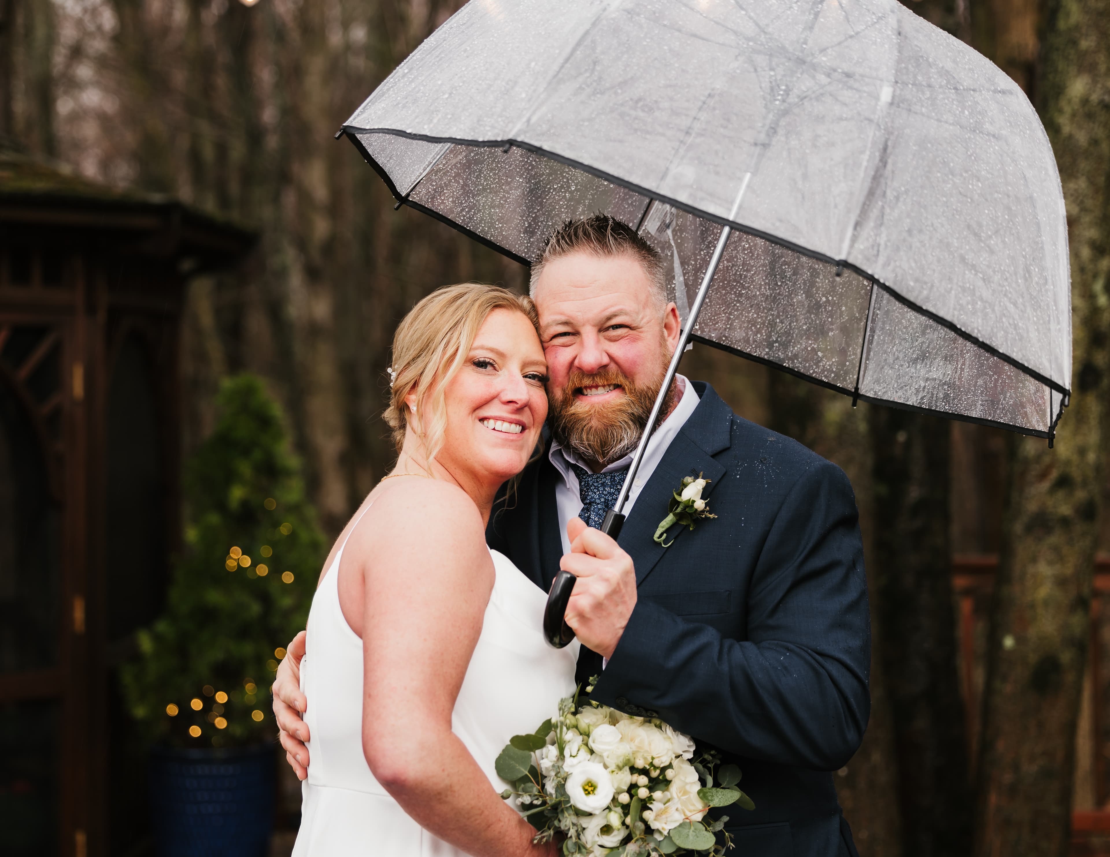 A smiling couple stands under a clear umbrella, embracing in a rainy outdoor setting.