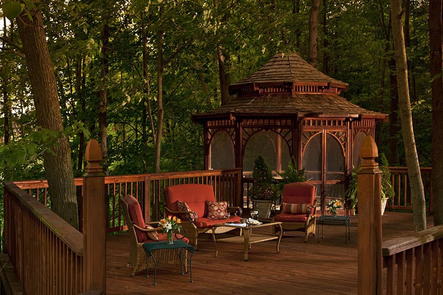 A cozy wooden deck with red chairs and a gazebo surrounded by lush trees.