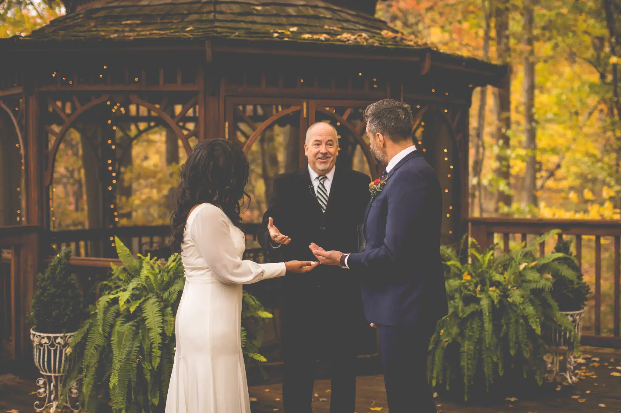 A couple stands in front of a officiant during an outdoor wedding ceremony, surrounded by greenery and autumn foliage.