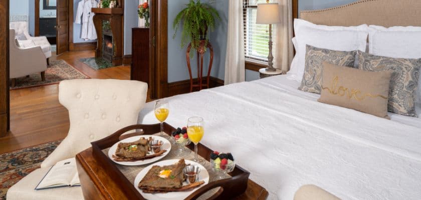 A cozy bedroom with a plush bed and a tray of breakfast items, including pancakes and drinks, on a chair in the foreground.