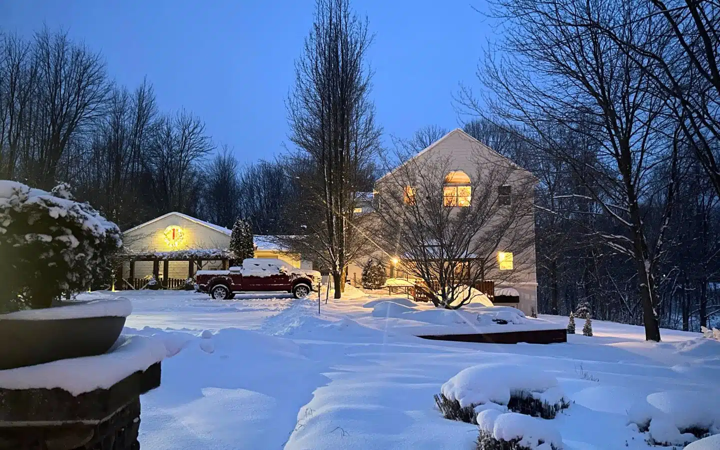 A snowy landscape features a two-story house with illuminated windows and a parked truck, set against a twilight sky.