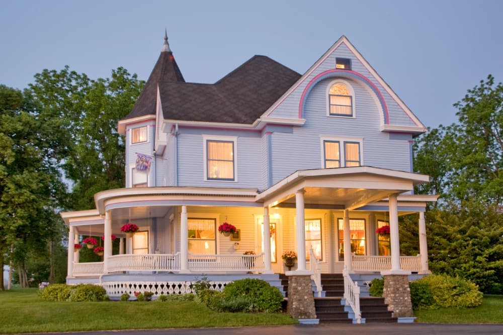 A charming blue Victorian-style house with a wraparound porch and colorful flower pots.