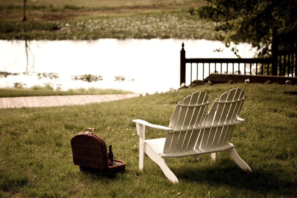 Two white Adirondack chairs sit on a grassy lawn near a calm pond, accompanied by a picnic basket and a bottle of wine.