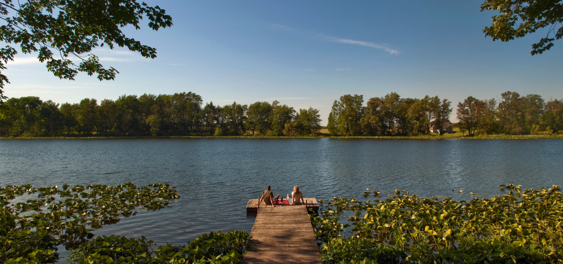 Two people sit on a wooden dock by a calm lake surrounded by lush greenery under a clear blue sky.