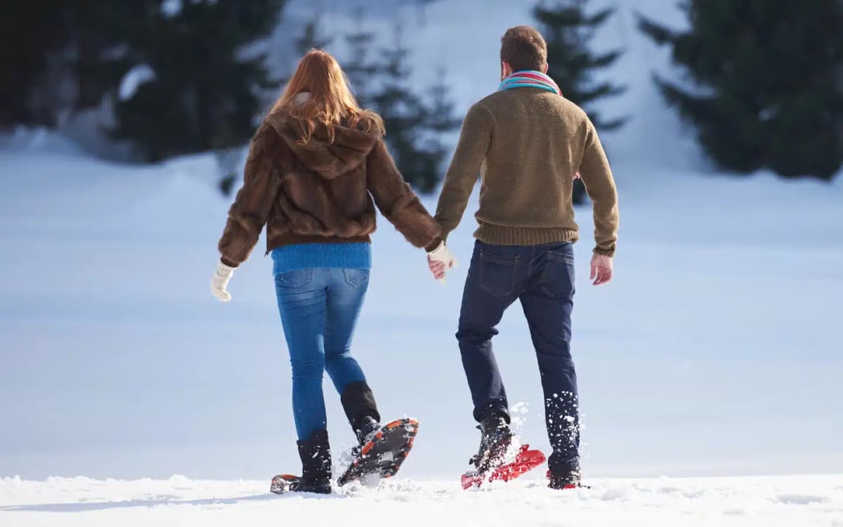 Man and woman holding hands while walking with snow shoes in a snow-covered field surrounded by evergreens