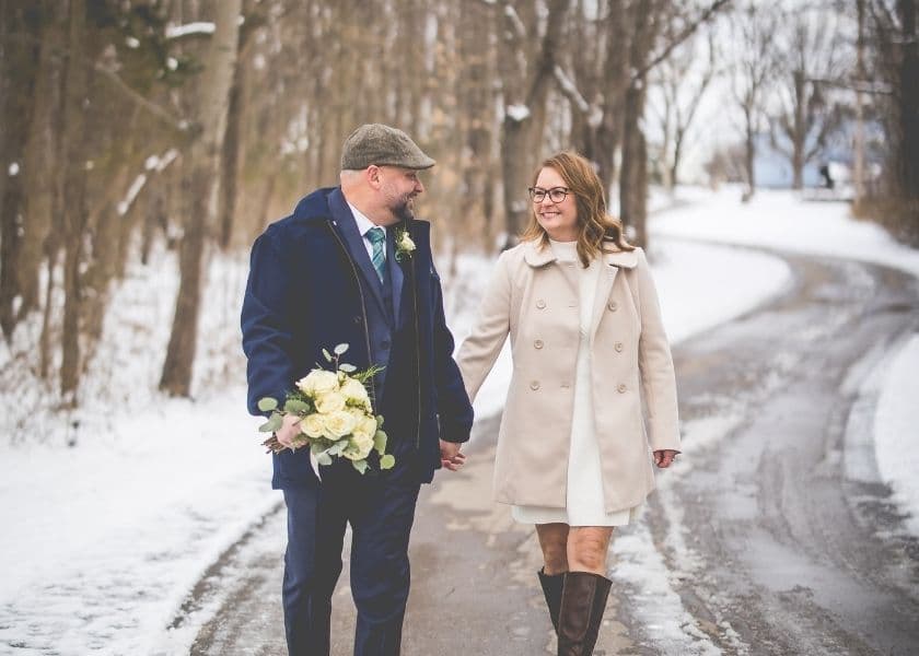 bridal couple walking down the snowy drive from castle in the country with the groom holding white roses in one hand and looking back at smiling bride bridal couple walking down the snowy drive from castle in the country with the groom holding white roses in one hand and looking back at smiling bride