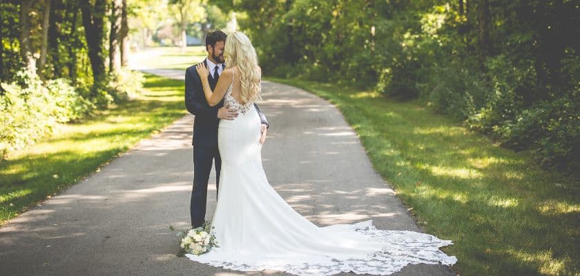 summer bridal couple embracing on the tree-lined driveway to castle in the country with white roses at the brides feet on her train